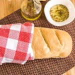 Golden loaf of French bread wrapped in a red and white towel beside a bowl of herb dipping oil.