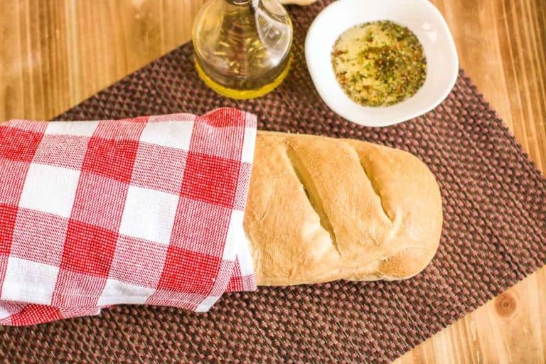 Golden loaf of French bread wrapped in a red and white towel beside a bowl of herb dipping oil.