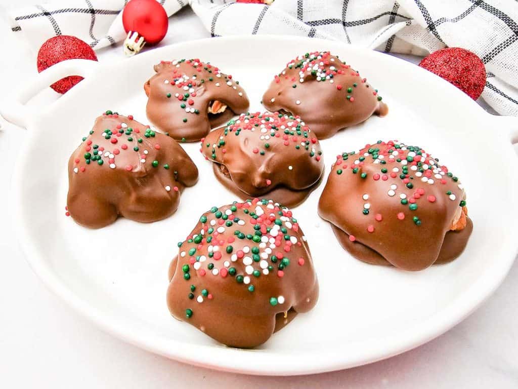 Angled view of six caramel pecan clusters coated in chocolate and decorated with holiday sprinkles, on a white plate.