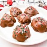 Angled view of six caramel pecan clusters coated in chocolate and decorated with holiday sprinkles, on a white plate.