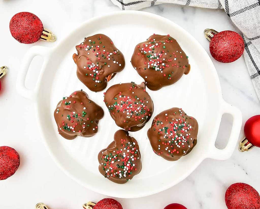 Six finished caramel pecan clusters coated in chocolate and topped with red, white, and green sprinkles on a white plate.