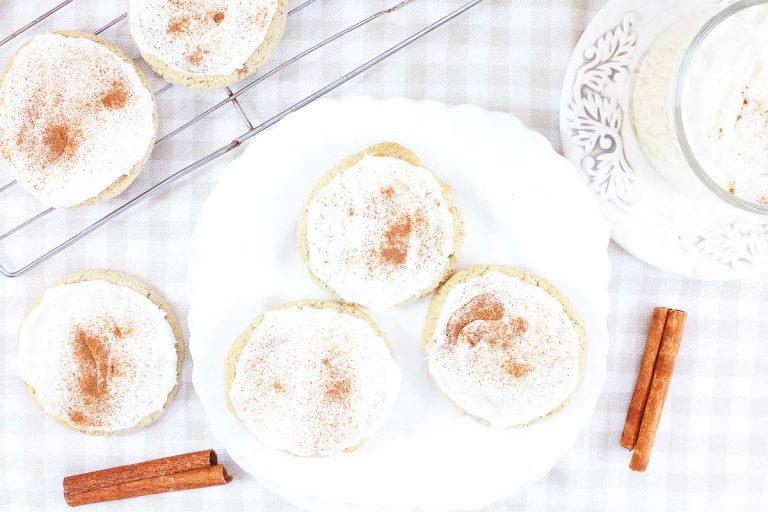A frosted eggnog cookie leaning against a mug of eggnog topped with whipped cream and cinnamon.