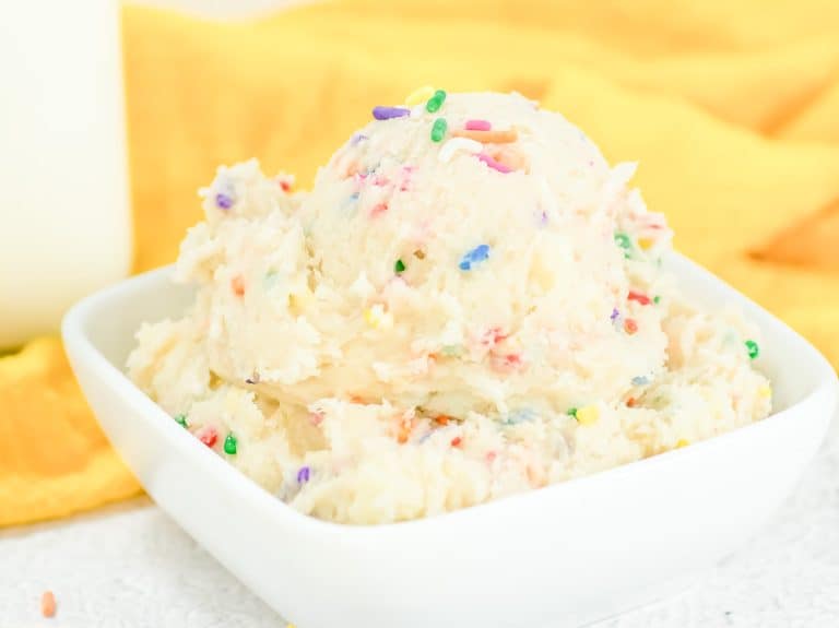 Bowl of creamy Funfetti edible cookie dough topped with colorful rainbow sprinkles, set against a yellow cloth.