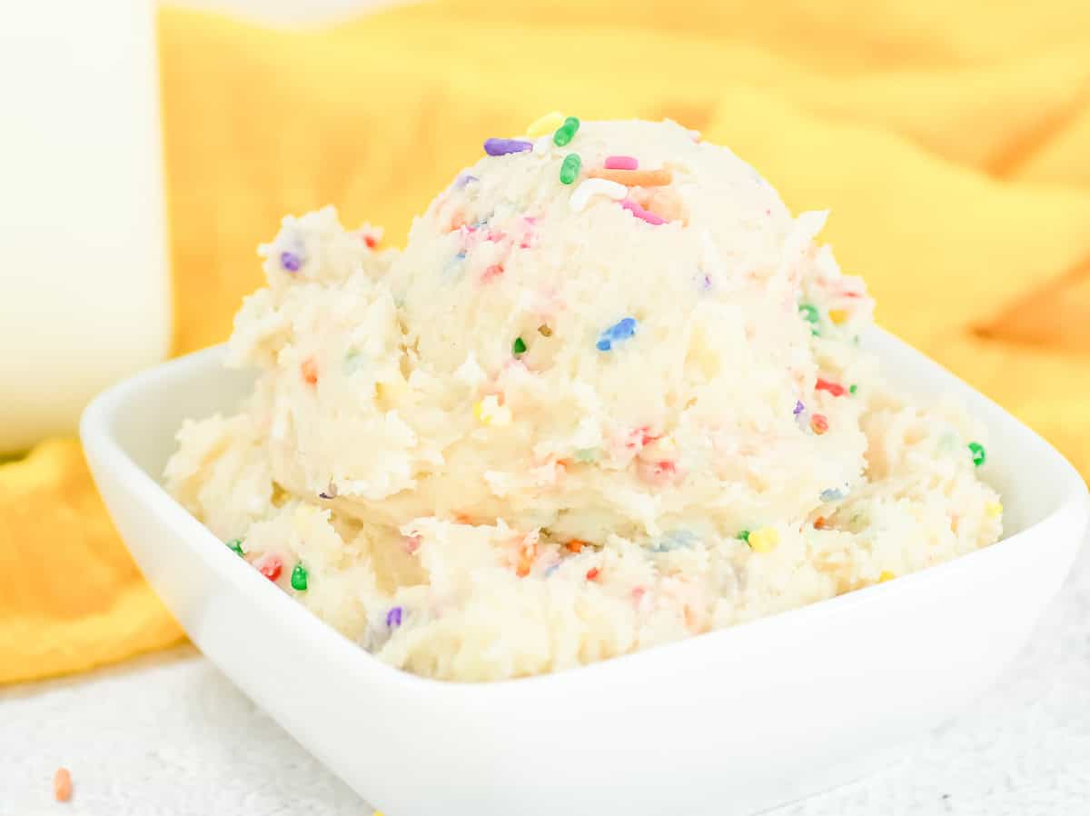 Bowl of creamy Funfetti edible cookie dough topped with colorful rainbow sprinkles, set against a yellow cloth.
