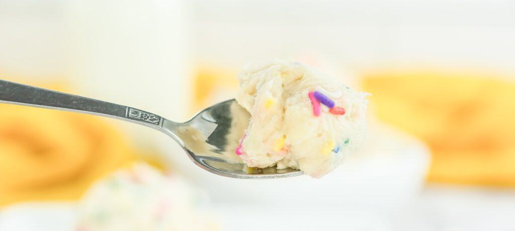 Spoon holding a bite of Funfetti edible cookie dough with rainbow sprinkles, lifted above a plate.