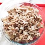 Glass bowl filled with white chocolate Chex mix on a red surface.