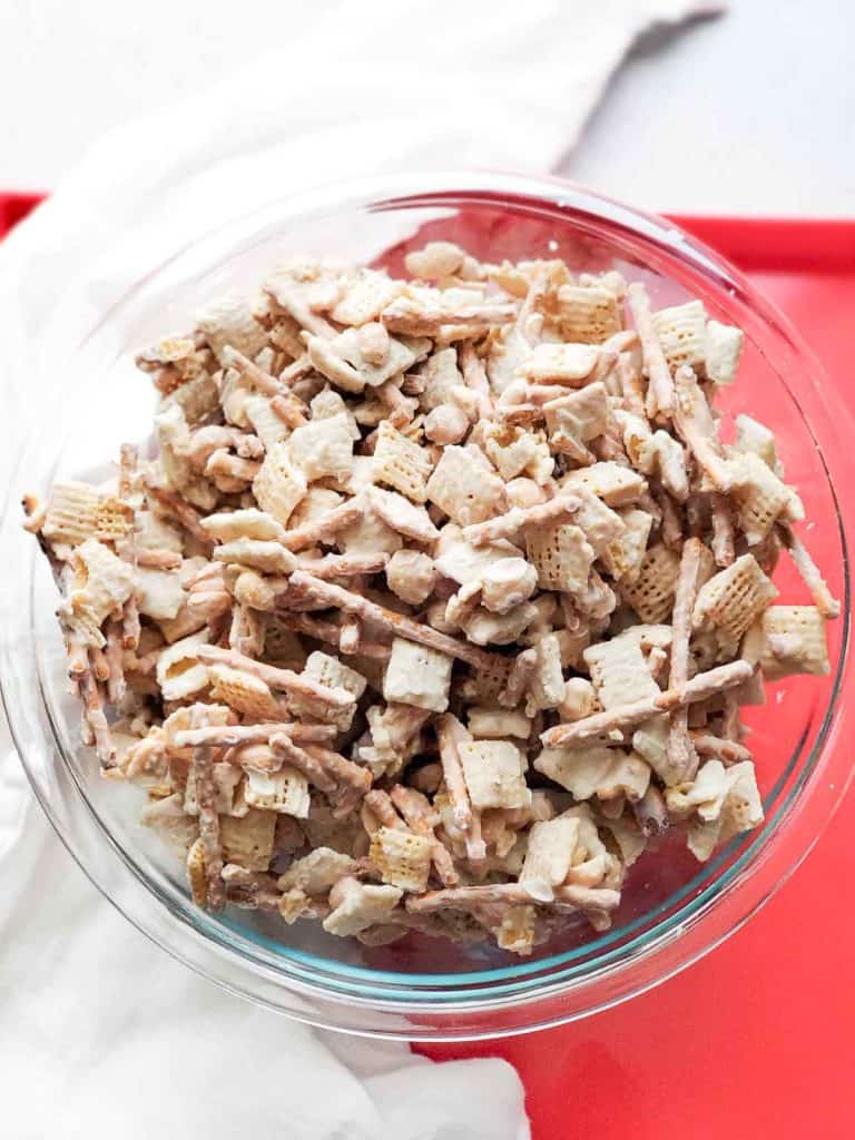 Glass bowl filled with white chocolate Chex mix on a red surface.