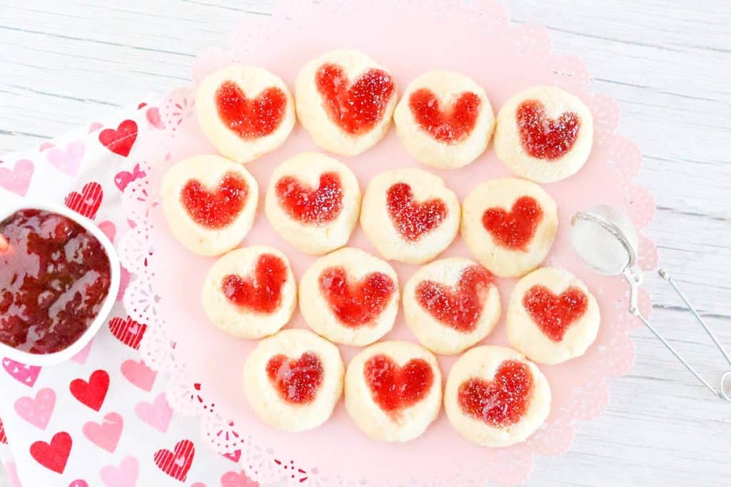 Finished heart thumbprint cookies on a pink doily-lined tray, lightly dusted with powdered sugar. A bowl of jam sits to the side on a heart-printed cloth.