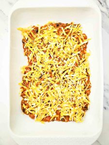 Shredded cheese added on top of enchilada casserole filling in a baking dish.