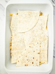 Flour tortillas layered over enchilada casserole with cheese in a baking dish.