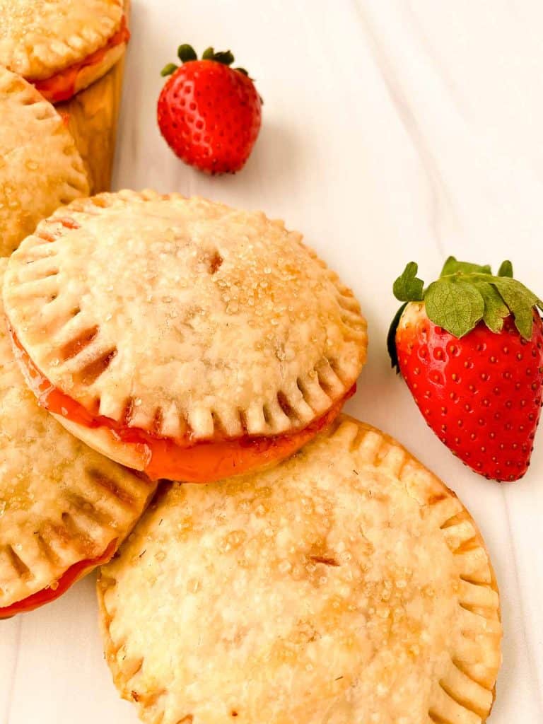 Close view of strawberry cream cheese hand pies with golden crusts and coarse sugar, with fresh strawberries nearby.