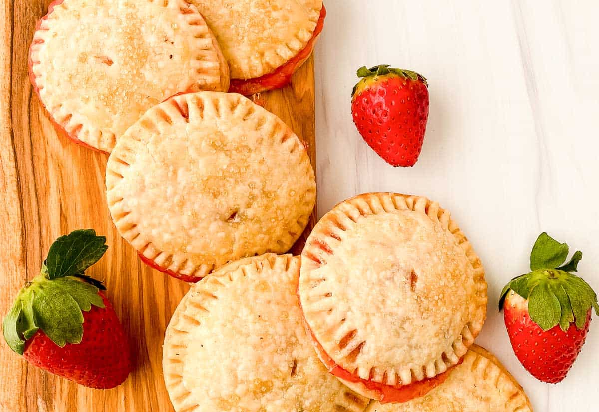 Strawberry cream cheese hand pies stacked on a wooden board with a fresh strawberry on the side.
