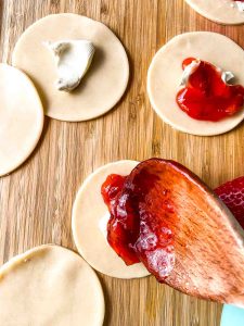 Pastry rounds on a cutting board topped with cream cheese and strawberry filling as a wooden spoon adds more filling.