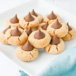 Close-up of peanut butter blossom cookies topped with chocolate kisses on a white plate, with a blue cloth nearby.