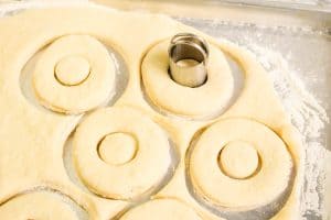 Donut shapes cut from rolled dough with a cutter, with centers removed and dough scraps around.