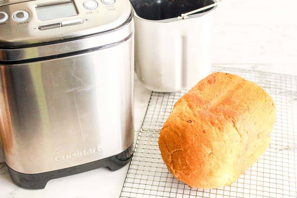 Freshly baked bread loaf cooling on a rack next to a bread machine.