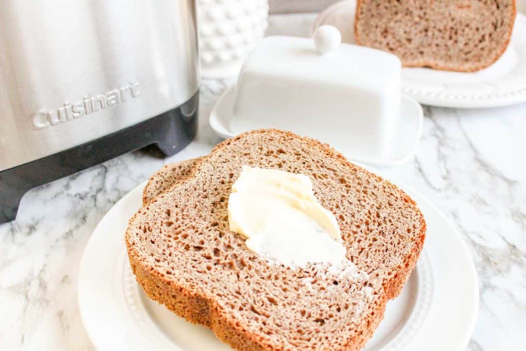 Slice of honey wheat bread topped with butter on a white plate beside a bread machine.